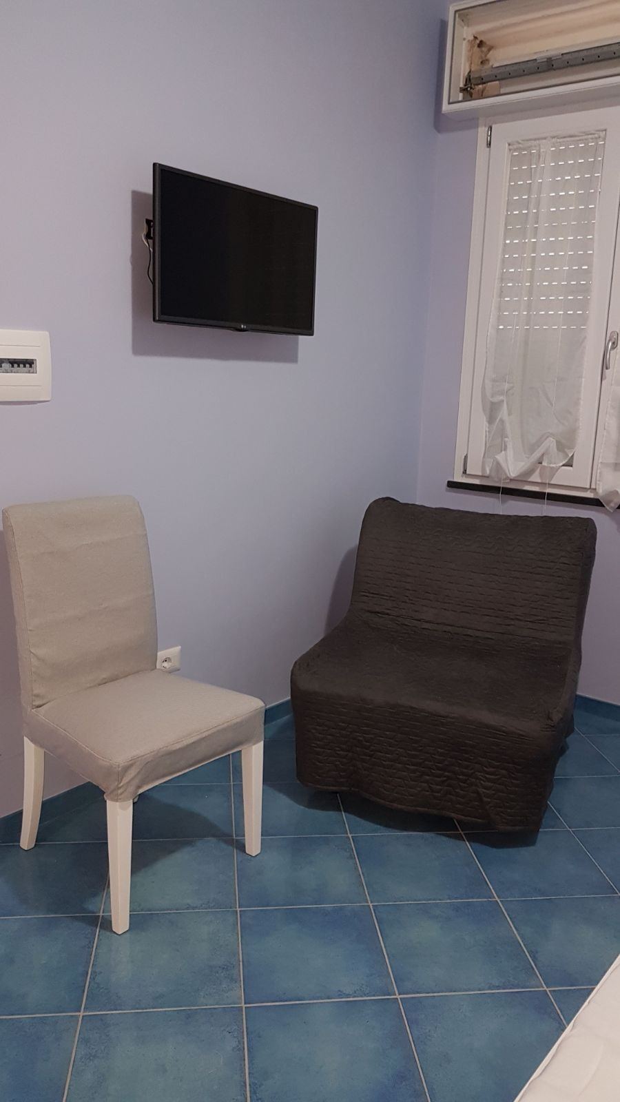 A modest room with lavender walls featuring a white dining chair, dark upholstered armchair, wall-mounted television, and blue tile flooring.