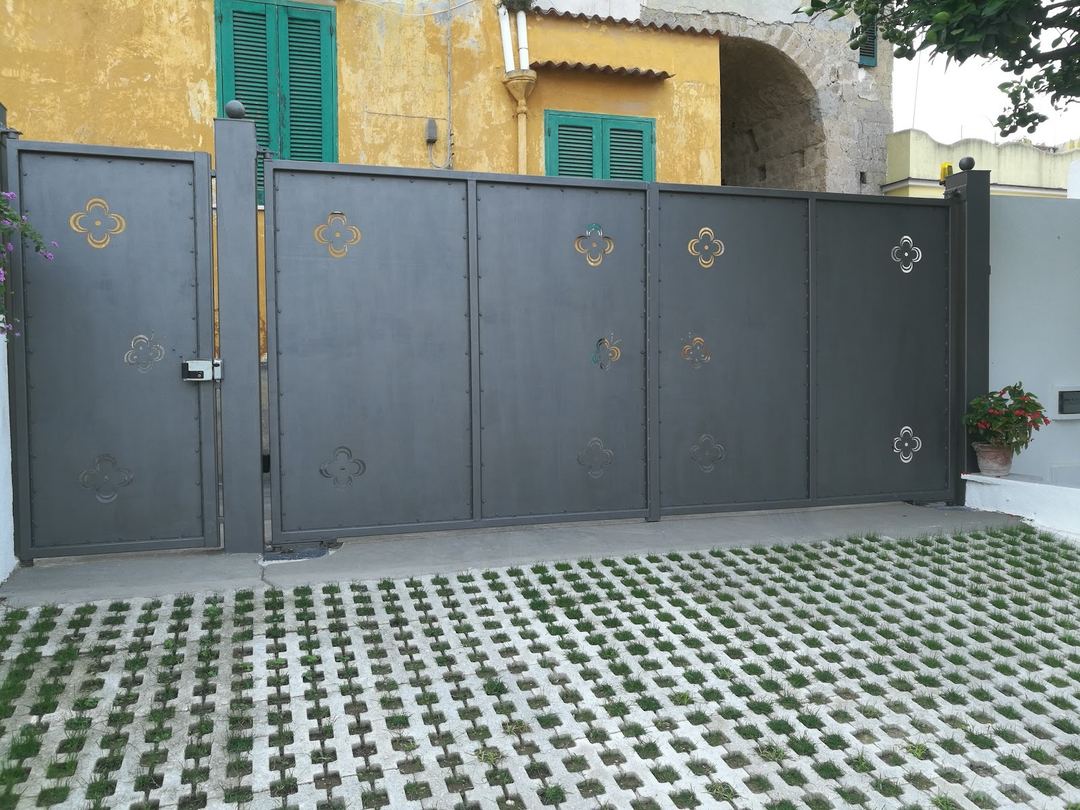 The entrance to the bed and breakfast features a dark metal gate with decorative motifs, permeable paving with grass, and a yellow ochre-colored building with green shutters.